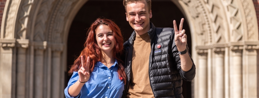 Rose Ritch stands on the left in a blue button-up shirt. Truman Fritz stands on the right in a dark khaki brown shirt and black jacket. Both are smiling as they throw up the “Fight On!” symbol. They stand in front of Bovard Auditorium on a sunny day.