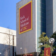 Photo of the Keck School of Medicine, a tall gray building with a banner reading “Keck School of Medicine of USC.” There are stairs leading up to the building, surrounding foliage, another building, and a blue sky in the background.