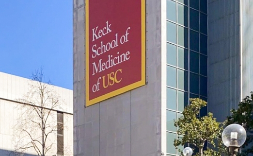 Photo of the Keck School of Medicine, a tall gray building with a banner reading “Keck School of Medicine of USC.” There are stairs leading up to the building, surrounding foliage, another building, and a blue sky in the background.