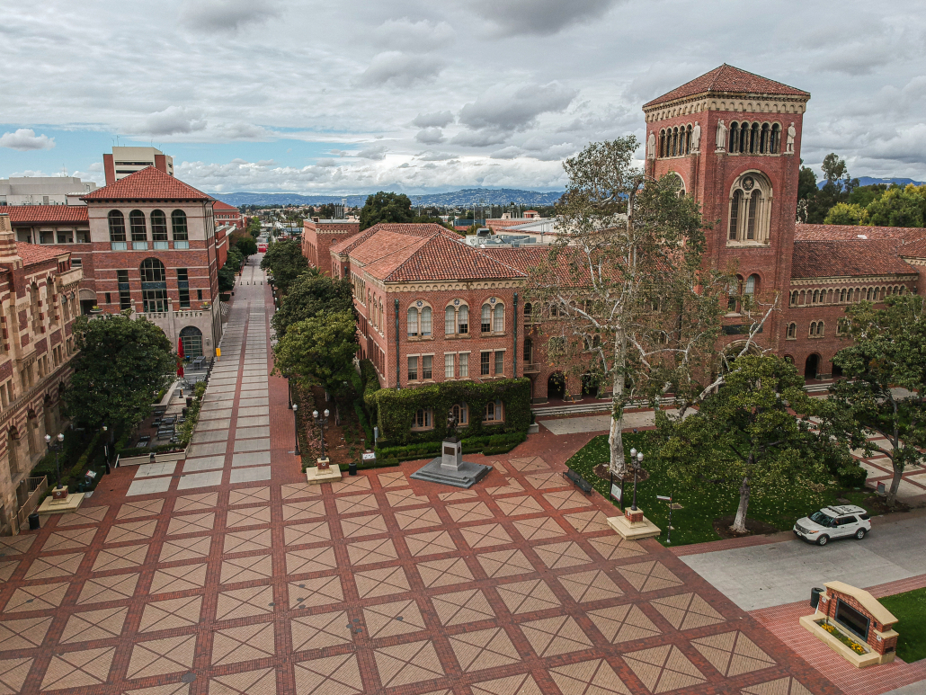 A partially gray-clouded sky overlooks the russet bricked buildings of Bovard Auditorium, the Student Union, Ronald Tutor Campus Center and the red and tan squared design of Hahn Plaza. A Department of Public Safety Cruiser sits in front of Bovard and the empty courtyard. 