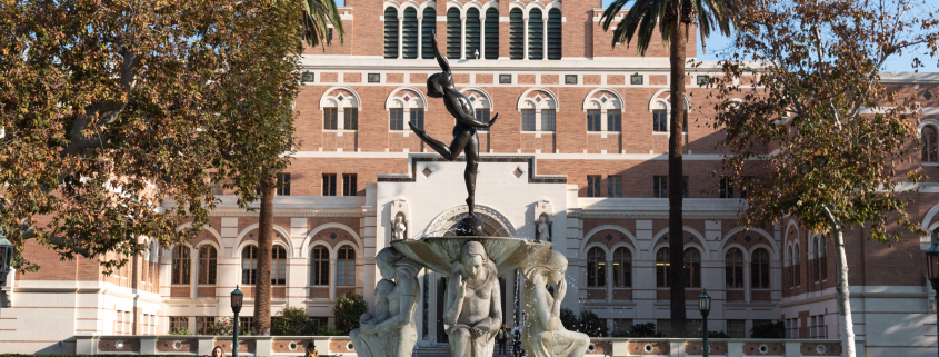 This is a photo of the fountain at the center of Alumni Park at USC. In the background is Doheny Memorial Library, partially in the shade on a bright sunny day.