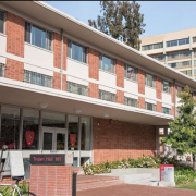 Photo of Trojan Hall residence hall, a red brick building with glass doors and windows and a sign in front reading “Trojan Hall 101.” There is another building, trees and blue sky in the background.