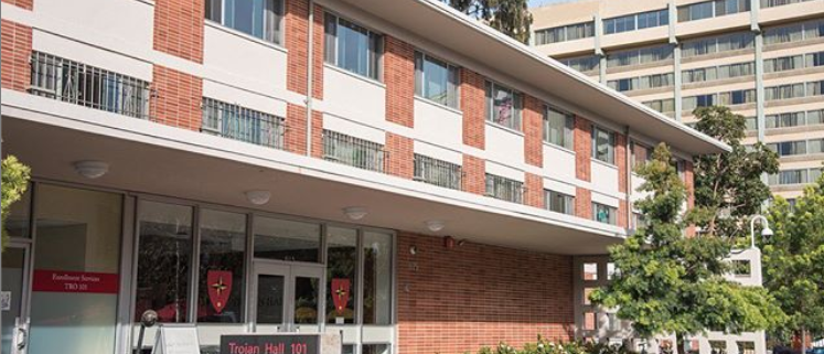 Photo of Trojan Hall residence hall, a red brick building with glass doors and windows and a sign in front reading “Trojan Hall 101.” There is another building, trees and blue sky in the background.