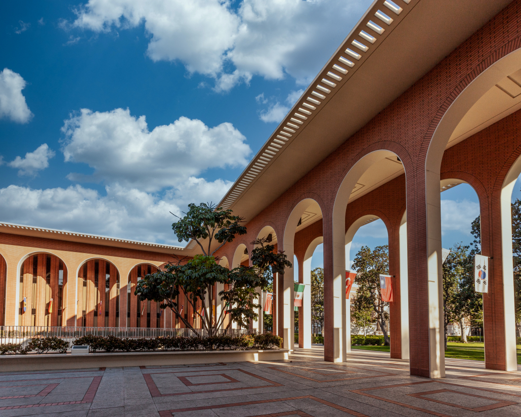 An arcuated red-bricked building, with its aisles partially lit and the courtyard shadowed by the partially exposed ceiling. Scattered white clouds and a blue sky overlook the building, with flags from various countries hang midway at the inner leg of each arch. 