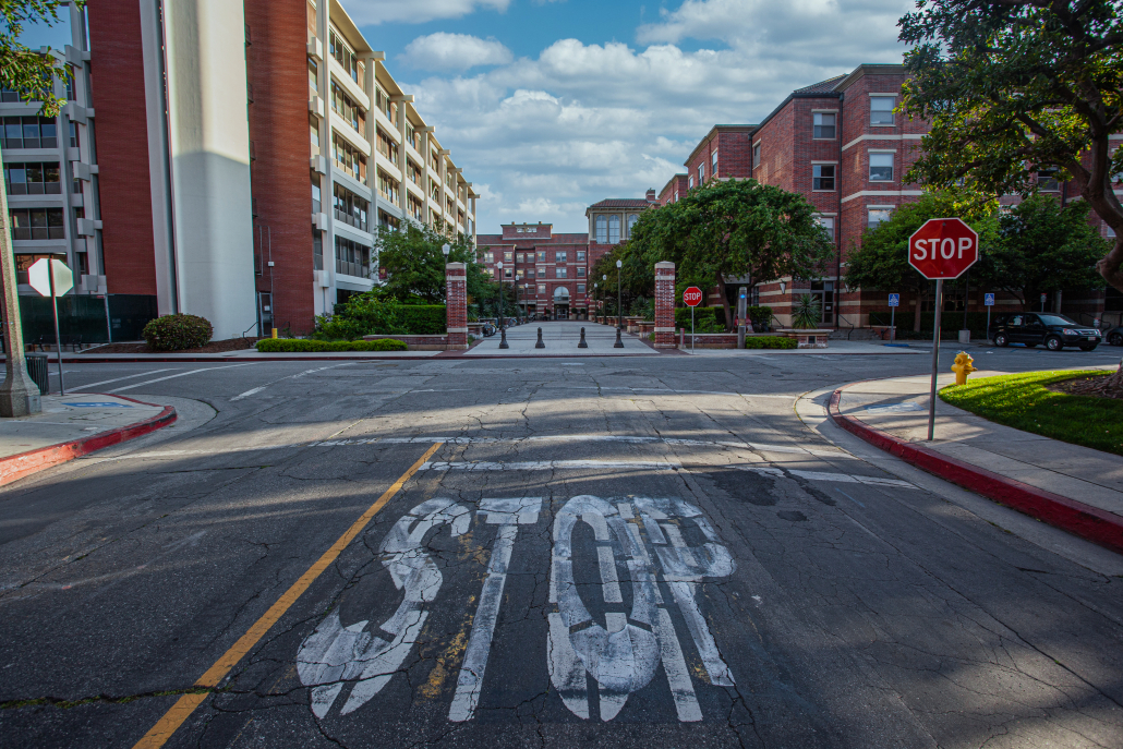 An overpainted white 'STOP' lays in the cracked and worn road leading into Parkside Apartments and Parkside dining hall. A vibrant red stop sign to the side of the road reiterates the white lettering and a white cloud scatter blue sky overlooks the buildings. 