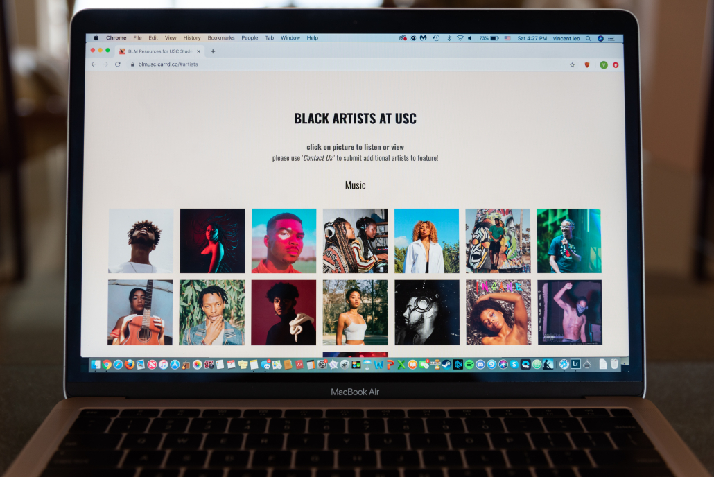 A Macbook Air screen displays a tab on the USC Black Lives Matter Resources page of “Black Artists AT USC,” with lettering at the top of the page. Two rows on the screen depict pictured squares of Black artists engaging in their medium or posing for the photo. 