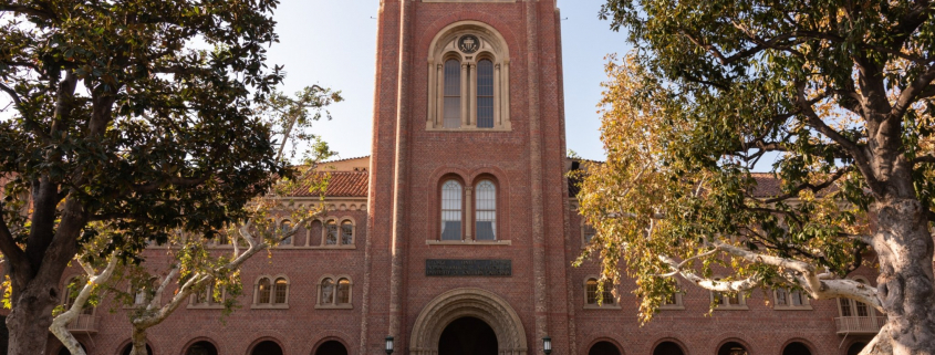 This is a wide angle photo of Board Auditorium on a bright sunny day. The center of Bovard is flanked by tall trees in Alumni Park.