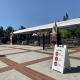 In front of one of the entrances to the campus, a COVID-19 check-in awaits visitors. There is also a white sign urging people to social distance, wear a mask and sanitize regularly.