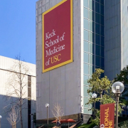 Photo of the Keck School of Medicine, a tall gray building with a banner reading “Keck School of Medicine of USC.” There are stairs leading up to the building, surrounding foliage, another building, and a blue sky in the background.