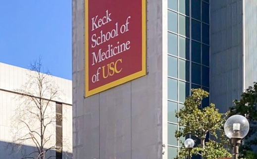 Photo of the Keck School of Medicine, a tall gray building with a banner reading “Keck School of Medicine of USC.” There are stairs leading up to the building, surrounding foliage, another building, and a blue sky in the background.