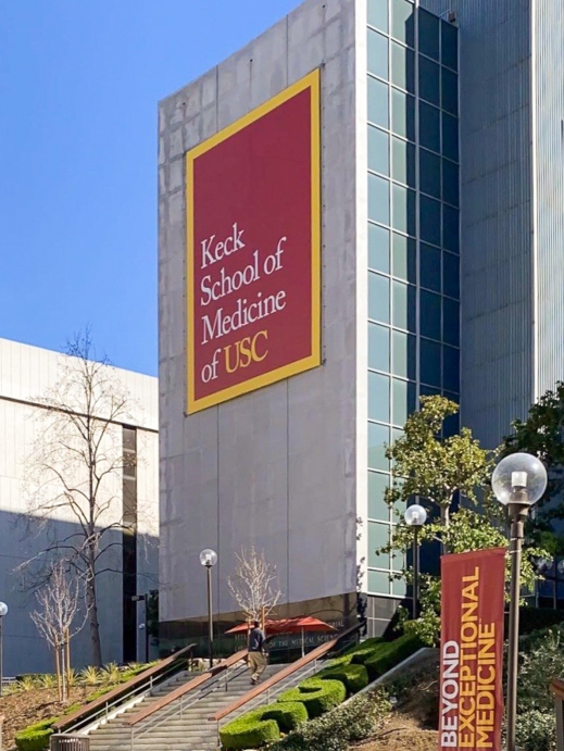 Photo of the Keck School of Medicine, a tall gray building with a banner reading “Keck School of Medicine of USC.” There are stairs leading up to the building, surrounding foliage, another building, and a blue sky in the background.