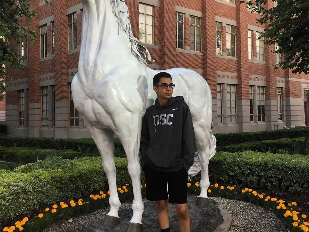 The photo shows Raymiro Gomez-Galiano standing next to USC’s mascot, Traveler, and is surrounded by a bed of yellow flowers. A three-story, red brick building with various glass windows sits in the background of Gomez-Galiano and Traveler. Gomez-Galiano also wears a grey USC shirt.