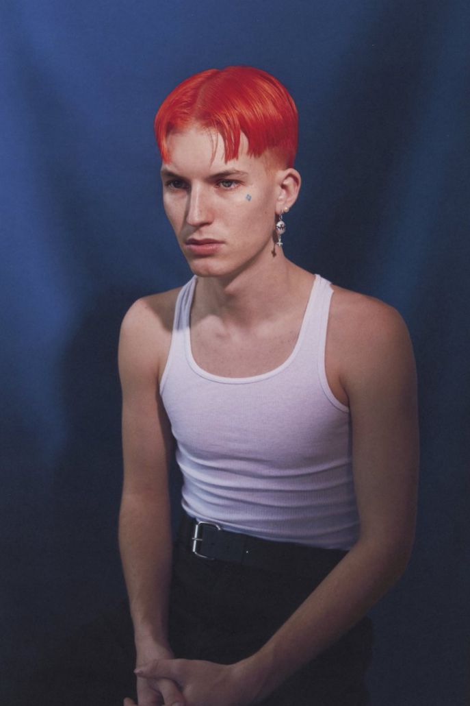 Gus Dapperton is sitting on a stool with black pants and a white tank top, with hair dyed orange-red and a somber expression on his face.