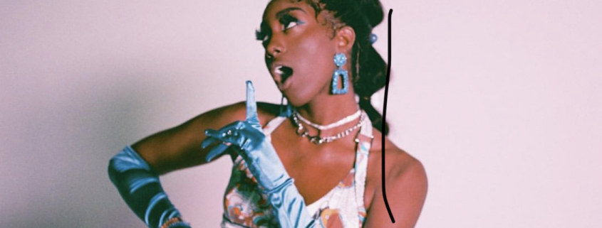 Rapper Flo Milli stands in front of a light pink background looking to her right. She dons baby blue gloves and matching dangling earrings. She's wearing a white printed dress and matching chokers and assorted necklaces. Her hair is pulled up into a long ponytail.
