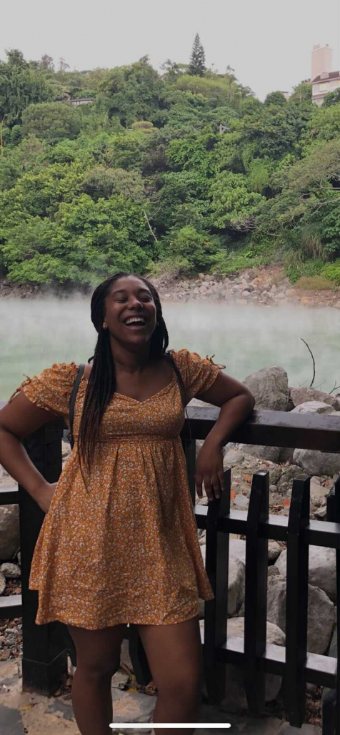 Photo of Jaya Hinton leaning on a brown, wooden fence. The picture /captures/ Hinton in mid-laugh with her eyes closed. In the background, a hill of various green-tinted trees lead into a bed of rocks, slightly hidden through a white mist of fog.