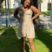 A photo of Jaya Hinton, smiling towards the camera with her hands on her hips. She wears a yellow and white checkered dress with an off-white building and palm trees in the background.