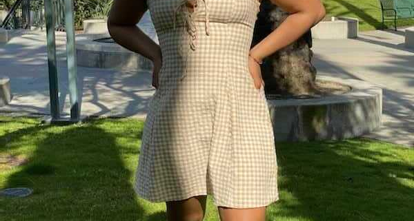 A photo of Jaya Hinton, smiling towards the camera with her hands on her hips. She wears a yellow and white checkered dress with an off-white building and palm trees in the background.