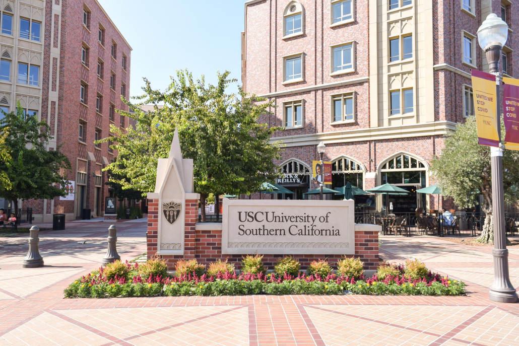 An image of the entrance to the USC Village with the restaurant Rock and Reilly’s in the background. There is a sign at the entrance that reads USC University of Southern California. On the right, there is a lamp post with flags that read “UNITED WE STOP COVID.”