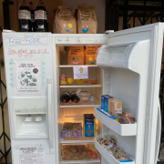 Photo of a stocked fridge with various food items including a variety of vegetables and bags of trail mix. On the closed fridge door marker scripted words read “Free Food!!” and “Take what you need.”