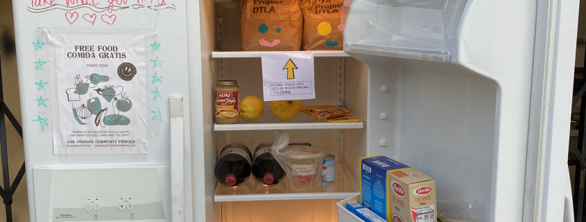 Photo of a stocked fridge with various food items including a variety of vegetables and bags of trail mix. On the closed fridge door marker scripted words read “Free Food!!” and “Take what you need.”