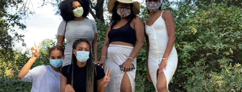 A group of five girls in front of trees with masks on. Three of them are standing at the back with two of them kneeling.