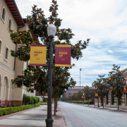 A sidewalk on campus with banners on lampposts telling people to "Mask up". On the left is the yellow building of the school of cinematic arts and next to the road is the red building of the uytengsu aquatic center
