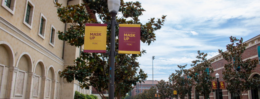 A sidewalk on campus with banners on lampposts telling people to "Mask up". On the left is the yellow building of the school of cinematic arts and next to the road is the red building of the uytengsu aquatic center