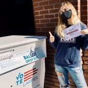 Emily Drysdale standing in front of a red brick wall next to a ballot drop box holding a mail-in ballot in her hand