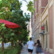 Backs of two people walking in the USC village with red brick building to their left and plants and trees to the right.