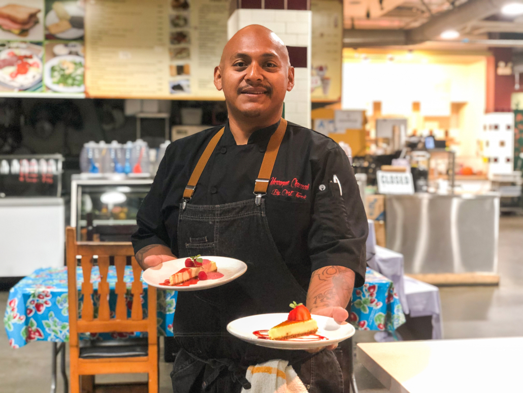 Chef Turok stands in front of a counter. He is wearing a black chef's uniform and is holding out two plates of cheesecake. 