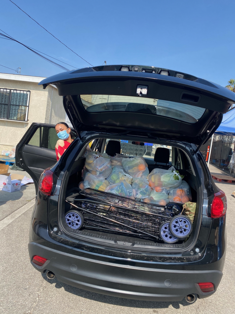 Volunteer stands next to car with open trunk, which is full of bags of food. 
