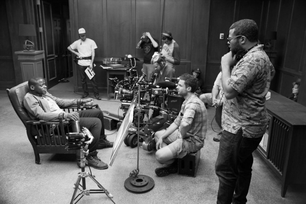 Jordan Peele sits on a chair with crew of filmmakers surrounding him. 