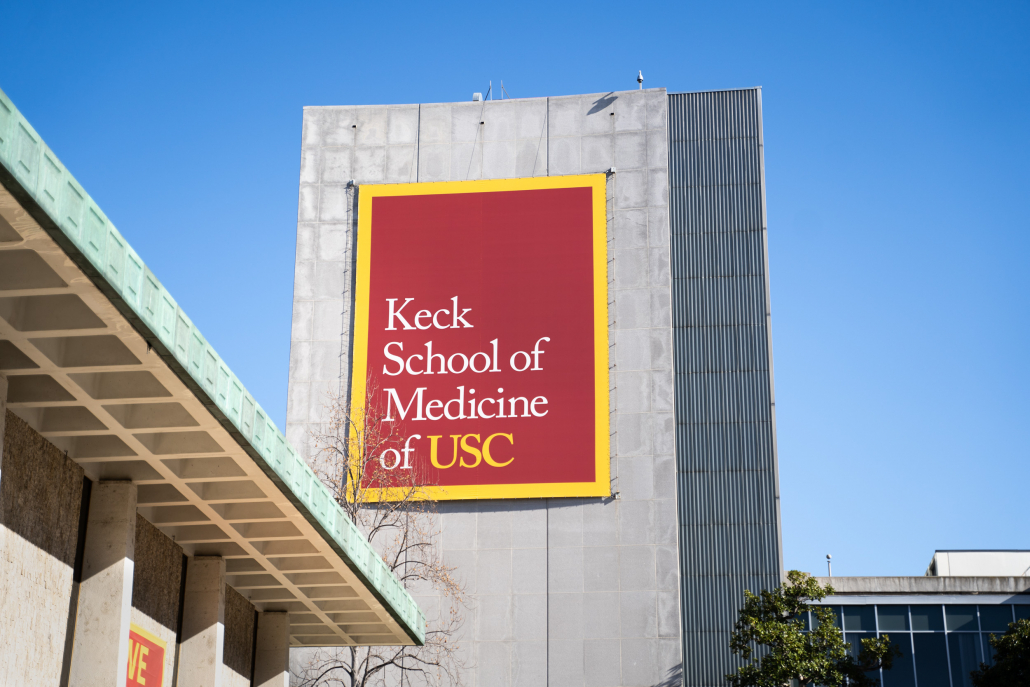 Photo of the Keck School of Medicine, a tall gray building with a banner reading “Keck School of Medicine of USC.” There are smaller buildings to the left and the right and the sky is blue.