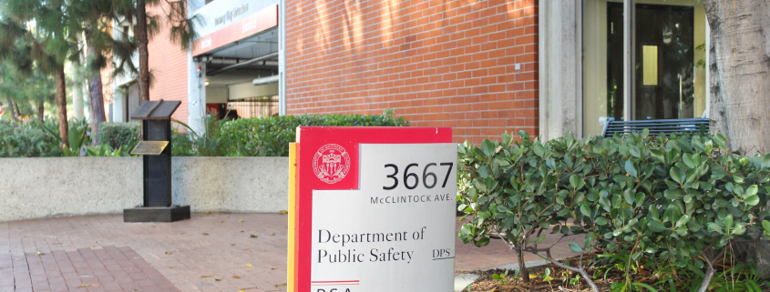 Photo of a red and grey sign that reads “Department of Public Safety.” Greenery and a red brick building connected to a windowed room surround the sign.