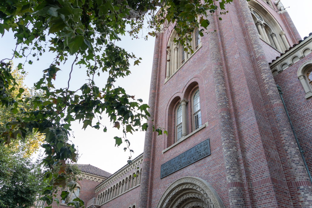 A low angle image of a various windowed tower on a red and brown bricked building. Tree branches and leaves surround the frame. 
