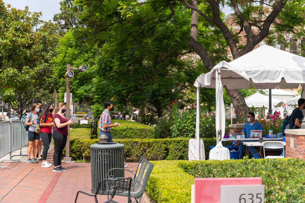 USC students stand in a line in front of the Pardee/Marks testing site as they wait for a man in a blue PPE suit. The man in blue is sitting on a chair behind a table with COVID test vials underneath a white tent.