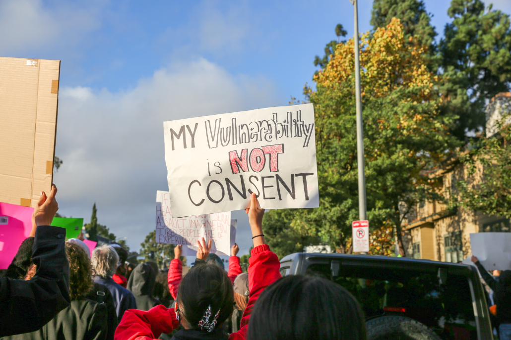 A photo of students marching and holding up signs. One sign reads "My vulnerability is not consent."