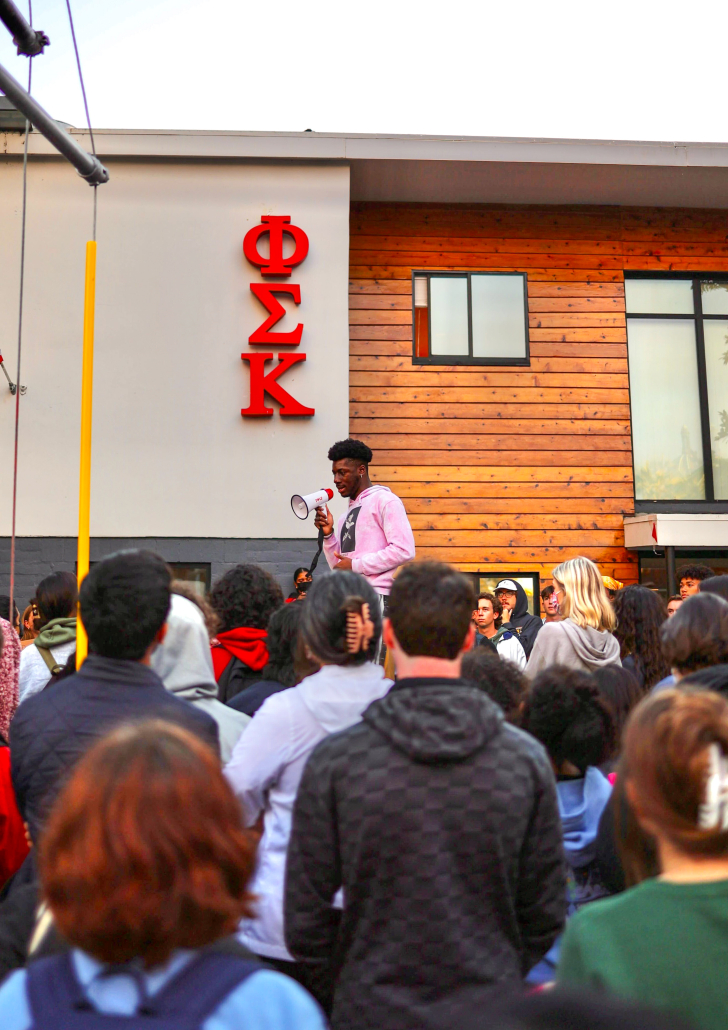 A photo of student speaking into a megaphone in front of the Phi Sigma Kappa fraternity house with other students gathered around him.