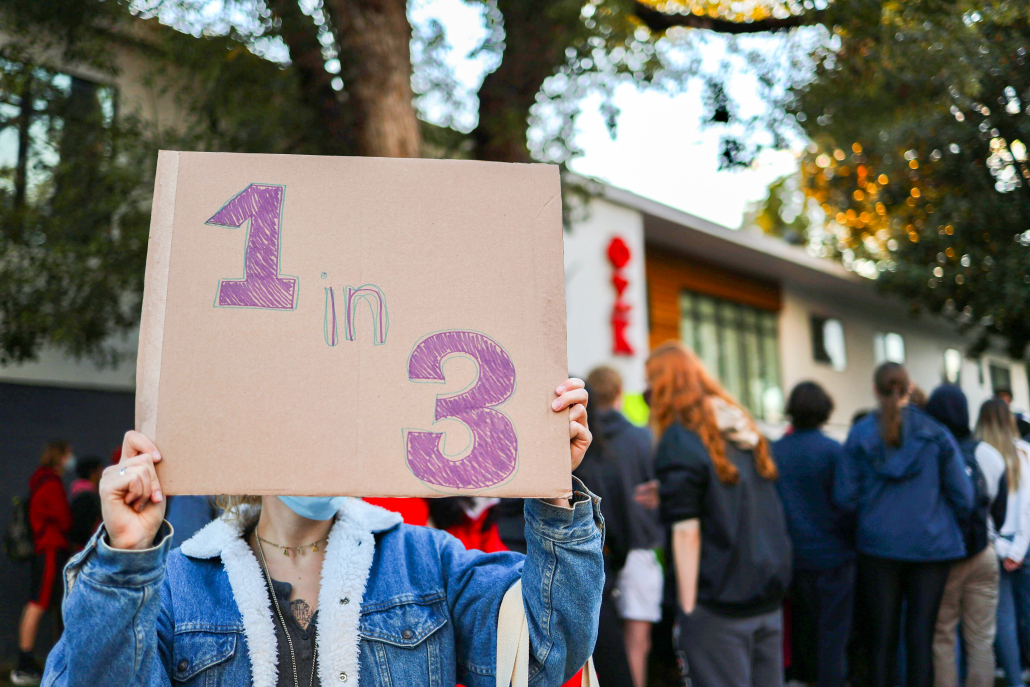 A photo of someone holding a sign that reads "1 in 3" in front of the Phi Sigma Kappa fraternity house.
