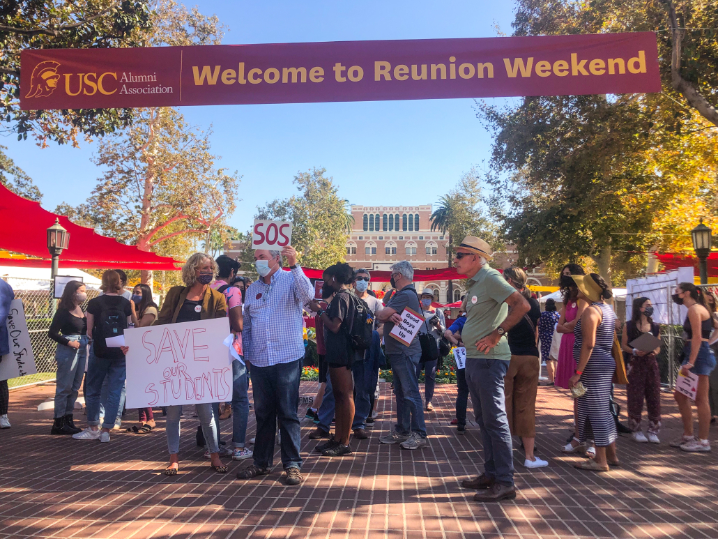 Protestors stand and hold signs outside Bovard. A banner stating "Welcome to Reunion Weekend" is held up by wires above thee protestors.