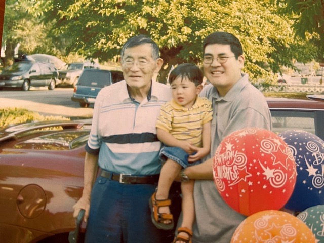 Image of the late Jiro Oishi with his grandson being held by his father. Behind them are trees and cars.