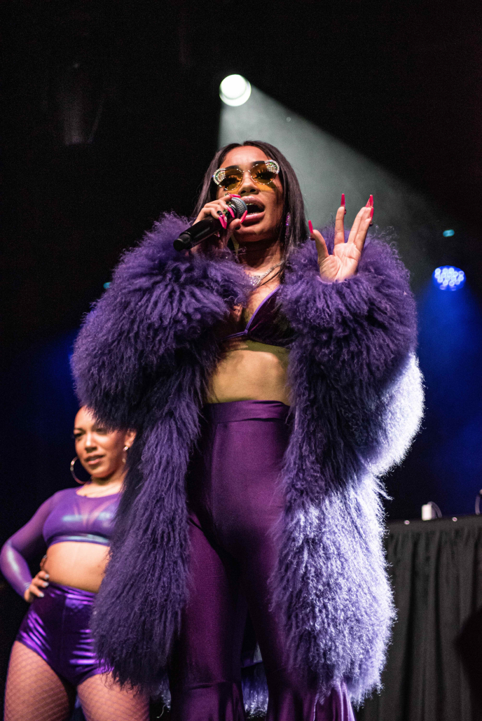 Photo of Saweetie. She is dressed in purple, has bright pink nails and has sunglasses on. There is a dancer in purple behind her.