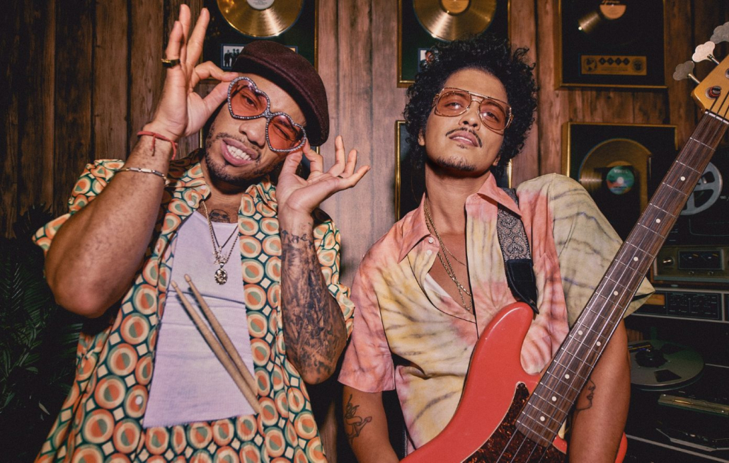 Two people in front of a wall of albums one holding sunglasses on their face and the other holding a bass guitar. 