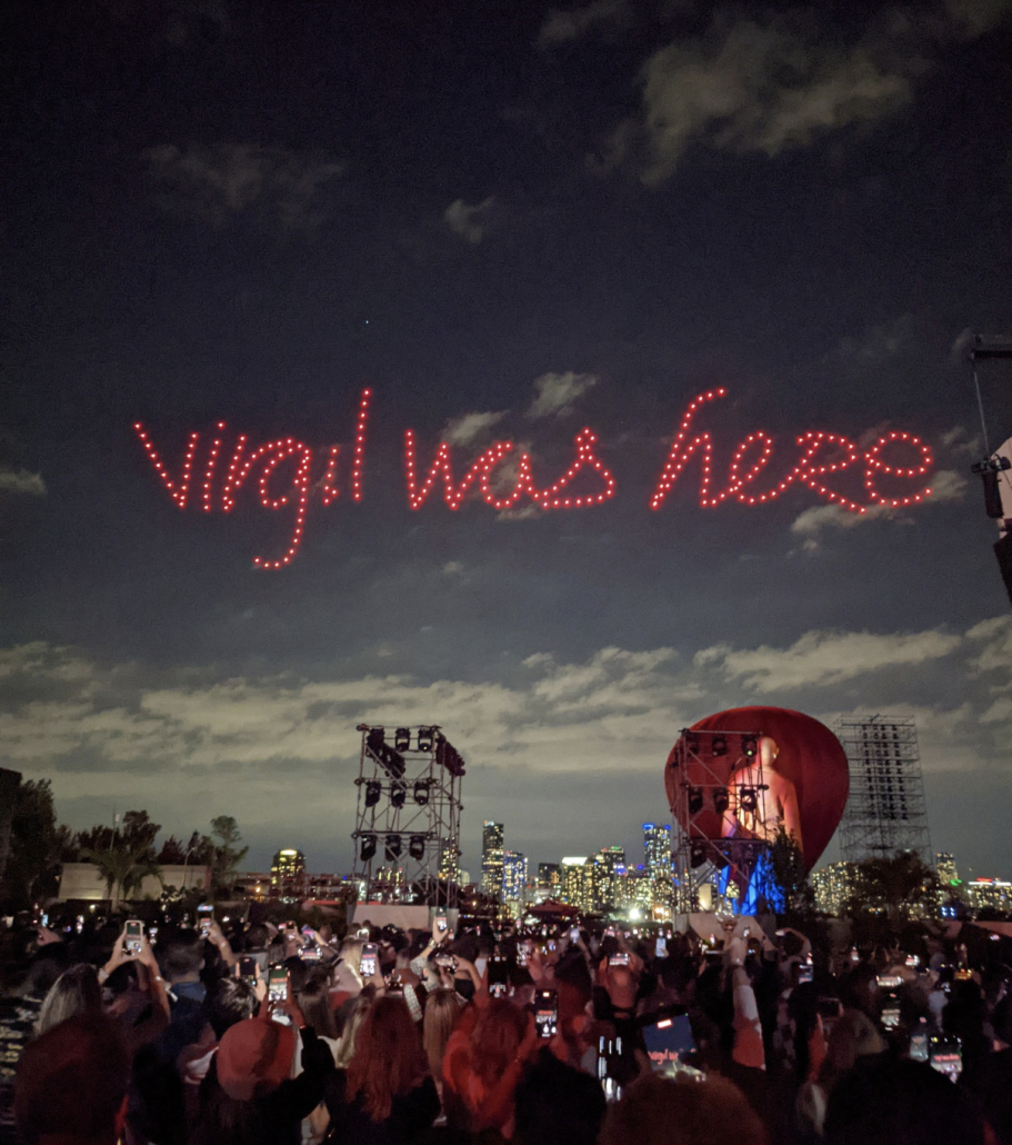 Photo of people at a concert. There is the words "virgil was here" in red lights in the sky.