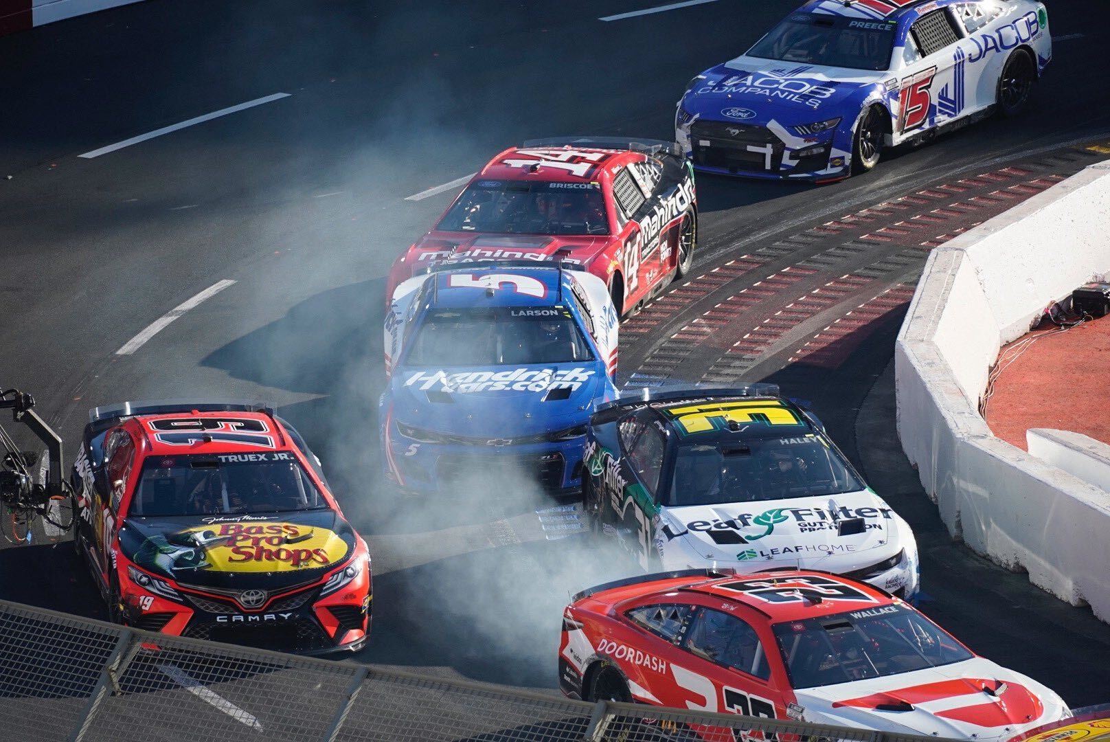 Race cars skid around the track, causing smoke to rise into the air. The racers are in the middle of a turn.