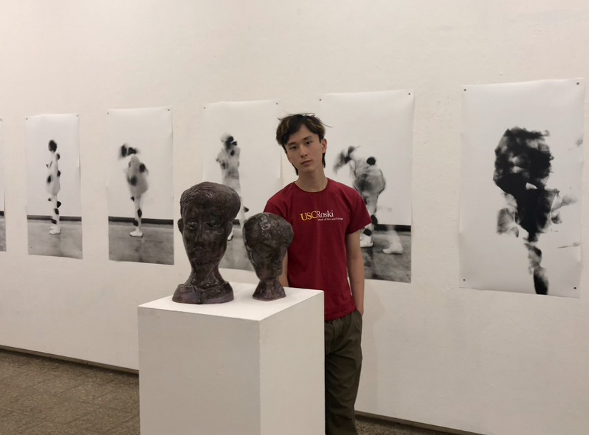 Brian Xu standing next to a two abstract sculptures of human heads. 