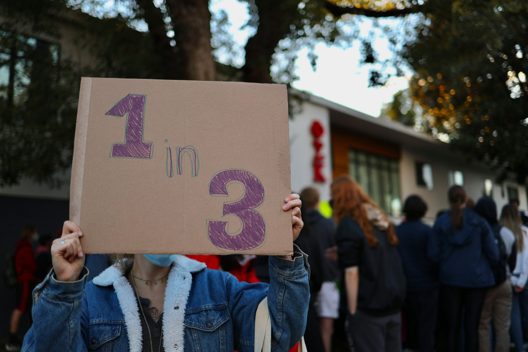 Photo of a girl at a protest holding a cardboard sign saying "1 in 3," in purple writing. 
