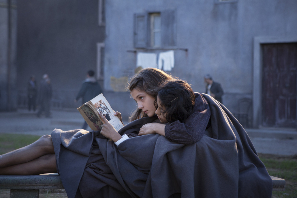 Two young girls read a book together on a bench, with one girl leaning against the other.