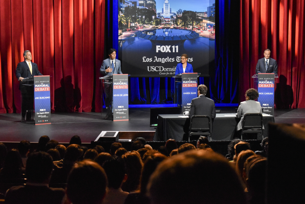 There are four candidates on the stage and the moderators are visible. The projection in the background reads FOX 11, Los Angeles Times and USC Dornsife. 