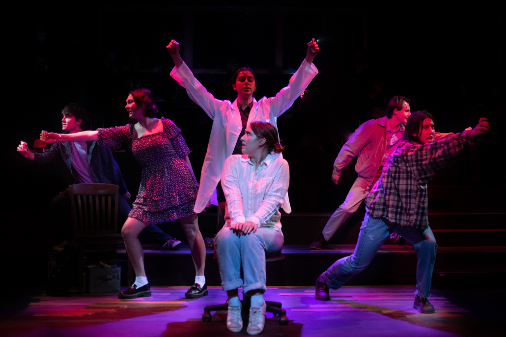 Still from the production of "next to normal." Still shows a woman sitting on stage looking surprised while people stand behind her pointing in different directions. 